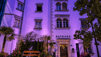 Das lila beleuchtete Rathaus in Spittal mit Blumenbeeten einer Bank und Bäumen im Vordergrund bei Nacht