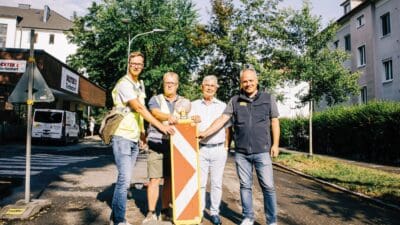 Vier Männer stehen auf einer Straße und halten gemeinsam ein Baustellenschild.