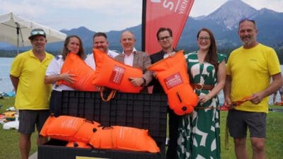 Sechs Personen präsentieren große orangene Leihbojen vor einem See mit Bergpanorama.