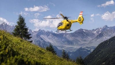Ein gelber Rettungshubschrauber fliegt über grüne Wiesen vor einer Bergkette mit Schnee