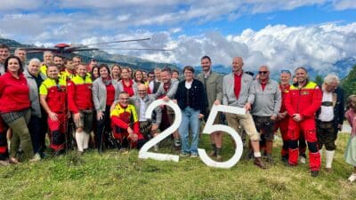 Große Gruppe von Rettungskräften und Gästen posiert mit weißer 25 vor Hubschrauber in Berglandschaft