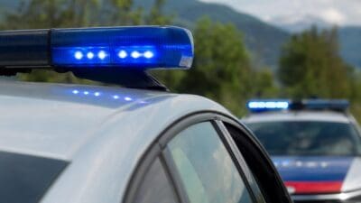 Nahaufnahme eines Polizeifahrzeugs mit blau leuchtendem Blaulichtbalken und einem zweiten Streifenwagen im Hintergrund vor Bergen.