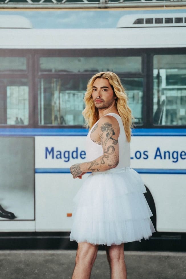 Tätowierte Person mit langen blonden Haaren in weißem Top und Tutu vor einem Bus
