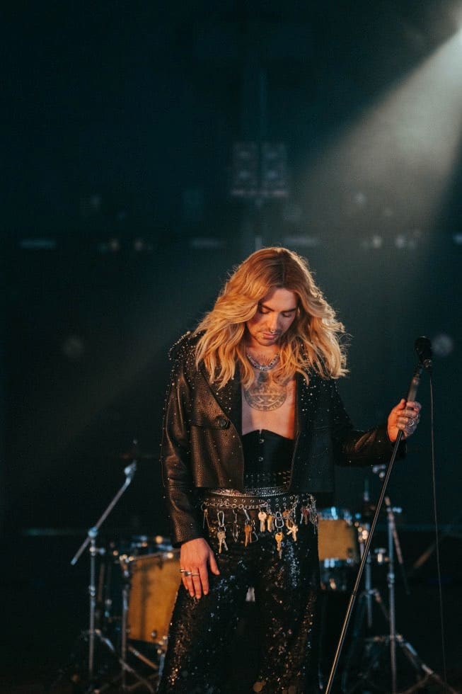 Männlicher Performer mit langen blonden Haaren in Lederjacke und Paillettenhose auf einer Bühne.