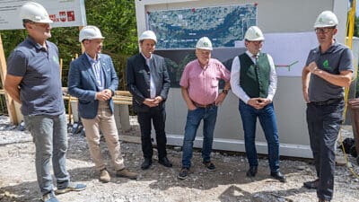 Sechs Männer mit weißen Schutzhelmen stehen vor einer großen Karte auf einer Baustelle.