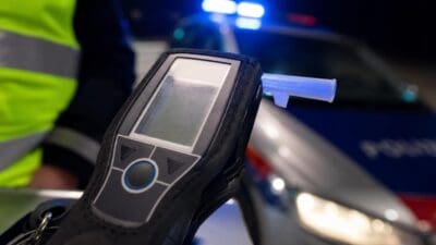 Eine Person in Warnweste hält ein Alkoholmessgerät vor einem Polizeifahrzeug mit Blaulicht