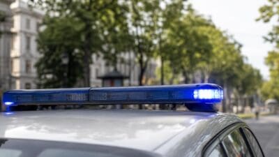 Blaues Blaulicht auf dem Dach eines silbernen Polizeifahrzeugs vor grünen Bäumen