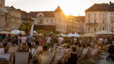 Menschen entspannen in weißen Liegestühlen auf einem sonnigen Platz in Klagenfurt mit historischen Gebäuden.