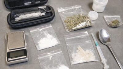 Plastikbeutel mit Cannabis und Kokain, digitale Waage, Löffel und Spritze auf grauem Untergrund