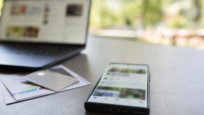 Ein Smartphone zeigt eine App mit Bildern auf einem Tisch neben einem Laptop und Karten.