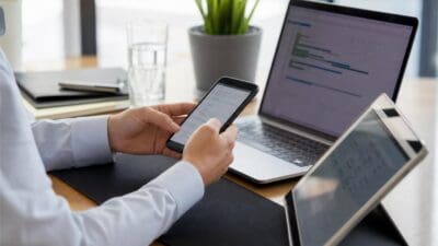 Eine Person in weißem Hemd bedient ein Smartphone vor einem Laptop und Tablet auf einem Schreibtisch.