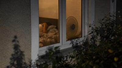 Durch ein Fenster ist eine schlafende Person in einem Bett mit einem Ventilator zu sehen.