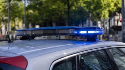 Aktiviertes blaues Blaulicht auf dem Dach eines silbernen Polizeifahrzeugs vor grünen Bäumen