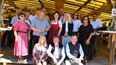Gruppe von Erwachsenen in Tracht und Alltagskleidung lächelt in die Kamera unter einem Holzdach.
