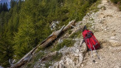 Ein roter Rucksack mit Wanderstöcken auf steinigem Pfad neben umgestürzten Baumstämmen im Bergwald