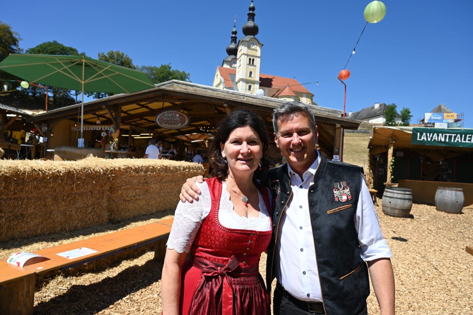 Eine Frau im roten Dirndl und ein Mann in Trachtenweste stehen vor einem Holzstand und einer Kirche.