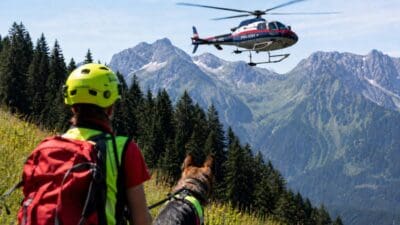 Person mit gelbem Helm und Hund beobachtet Polizeihubschrauber vor Bergpanorama