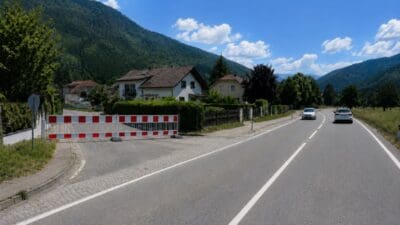 Rote und weiße Baustellenabsperrung blockiert eine Zufahrt neben einer Alpenstraße mit fahrenden Autos und Bergen im Hintergrund.