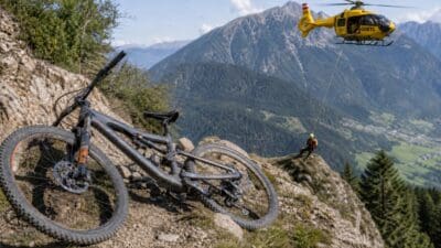 Graues Mountainbike auf steinigem Hang mit gelbem Rettungshubschrauber und Bergretter in alpiner Landschaft