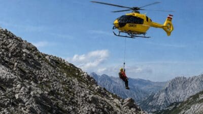 Ein gelber Rettungshubschrauber mit einem am Seil hängenden Retter über einem steinigen Berg in den Alpen.