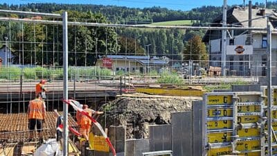 Mehrere Bauarbeiter in orangenen Warnwesten auf einer Baustelle hinter einem Zaun am Bahnhof Rothenthurn.