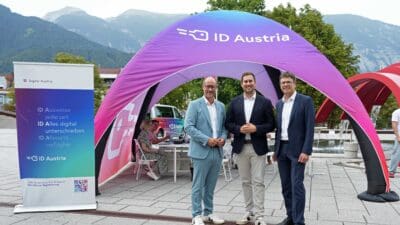 Drei Männer in Anzügen stehen vor einem großen lila ID Austria Zelt mit Bergen im Hintergrund.