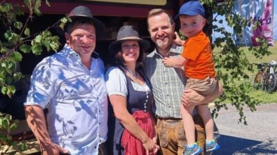 Vier Personen in traditioneller Tracht lächeln in die Kamera vor einem Fahrzeug auf einer Wiese.