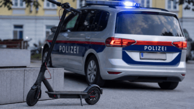 Ein schwarzer E-Scooter steht neben einem silbernen Polizeiauto mit eingeschaltetem Blaulicht.