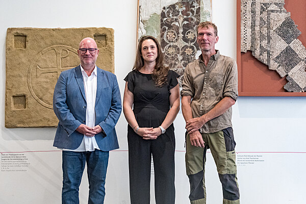 Drei Personen stehen vor antiken Steinreliefs und Mosaiken in einem Ausstellungsraum.