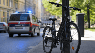 Schwarzes Fahrrad lehnt an einem dunklen Pfosten auf dem Gehweg vor einem Polizeiwagen mit Blaulicht