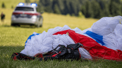 Zusammengefallener Gleitschirm in Rot, Weiß und Blau auf grüner Wiese mit Polizeiauto und Blaulicht im Hintergrund.