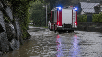 Feuerwehrfahrzeug mit Blaulicht fährt durch Hochwasser auf einer Straße im Regen.