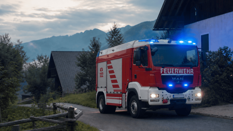 Rotes Feuerwehrfahrzeug mit blauem Blaulicht auf einer Landstraße vor Bergen bei Abenddämmerung