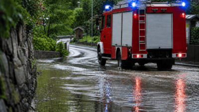 Rotes Feuerwehrfahrzeug mit aktiviertem Blaulicht fährt durch eine überflutete Straße.