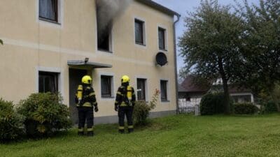Zwei Feuerwehrleute in Schutzkleidung und gelben Helmen stehen vor einem gelben Wohnhaus mit Rauch aus einem Fenster.
