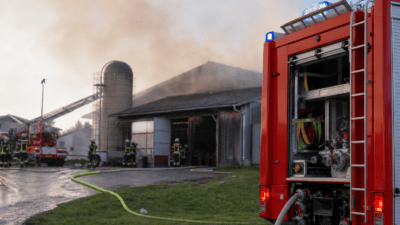 Rotes Feuerwehrfahrzeug mit offener Seite Schläuchen und Blaulicht vor rauchendem Wirtschaftsgebäude und Einsatzkräften.