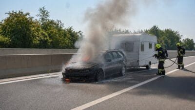 Zwei Feuerwehrleute löschen brennenden Pkw mit Wohnwagen auf der Autobahn