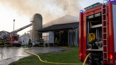 Feuerwehrleute in Schutzkleidung und rote Einsatzfahrzeuge vor einer verrauchten Scheune mit Silo
