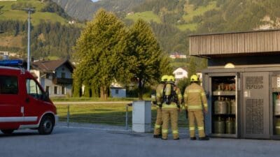 Drei Feuerwehrleute in Schutzausrüstung stehen vor einem geöffneten Gasflaschenautomaten neben einem roten Feuerwehrfahrzeug in bergiger Landschaft.