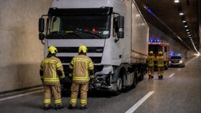 Zwei Feuerwehrleute in Schutzkleidung stehen vor der Front eines verunfallten weißen Lkw im Tunnel.