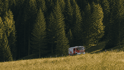 Rotes Feuerwehrfahrzeug parkt auf einer Wiese vor einem dichten Wald am Hang.