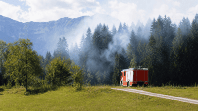 Rotes Feuerwehrfahrzeug steht auf einem Weg am Waldrand vor einem rauchenden Wald mit Bergen im Hintergrund