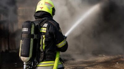 Rückansicht eines Feuerwehrmanns mit gelbem Helm und Atemschutz der einen Wasserstrahl in dichten Rauch sprüht