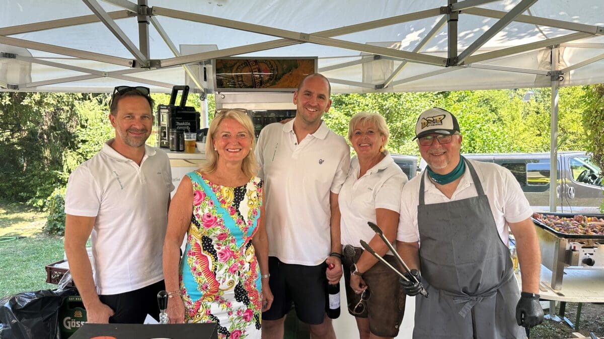 Fünf Erwachsene lächeln unter einem weißen Zelt beim Dorffest mit Getränken und Grill im Vordergrund.
