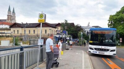 Weiß-blauer Bus mit Aufschrift Schienenersatzverkehr St. Andrä-Wördern fährt an Bushaltestelle mit wartenden Personen und Kinderwagen vorbei