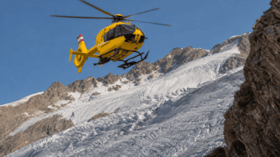 Gelber Hubschrauber fliegt über schneebedecktem Gletscher vor steilen Berggipfeln.