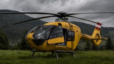 Gelber Hubschrauber mit geöffneten Türen auf einer Wiese vor Bergen