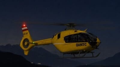 Gelber ÖAMTC-Rettungshubschrauber mit roter Leuchte fliegt bei Nacht über einer Berglandschaft.