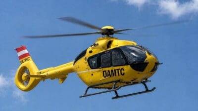 Gelber ÖAMTC-Rettungshubschrauber mit rot-weißem Heck und rotierenden Rotorblättern vor blauem Himmel.
