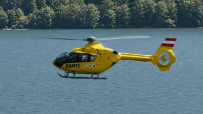 Gelber ÖAMTC-Rettungshubschrauber fliegt über einem See vor dichtem Wald am Ufer